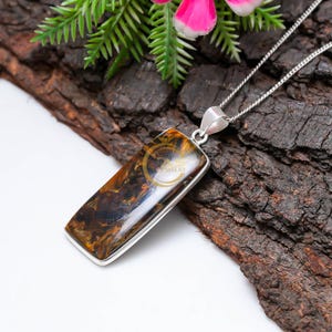 May include: A rectangular pendant with a silver-colored frame and chain. The pendant features a polished, brown and gold stone with a unique pattern. The jewelry is displayed on a textured, dark brown surface.