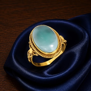 May include: A gold ring featuring a large, smooth, oval-shaped light blue stone set in a decorative twisted rope bezel. The band and setting include intricate filigree metalwork details, creating a polished and ornate appearance for this jewelry piece.