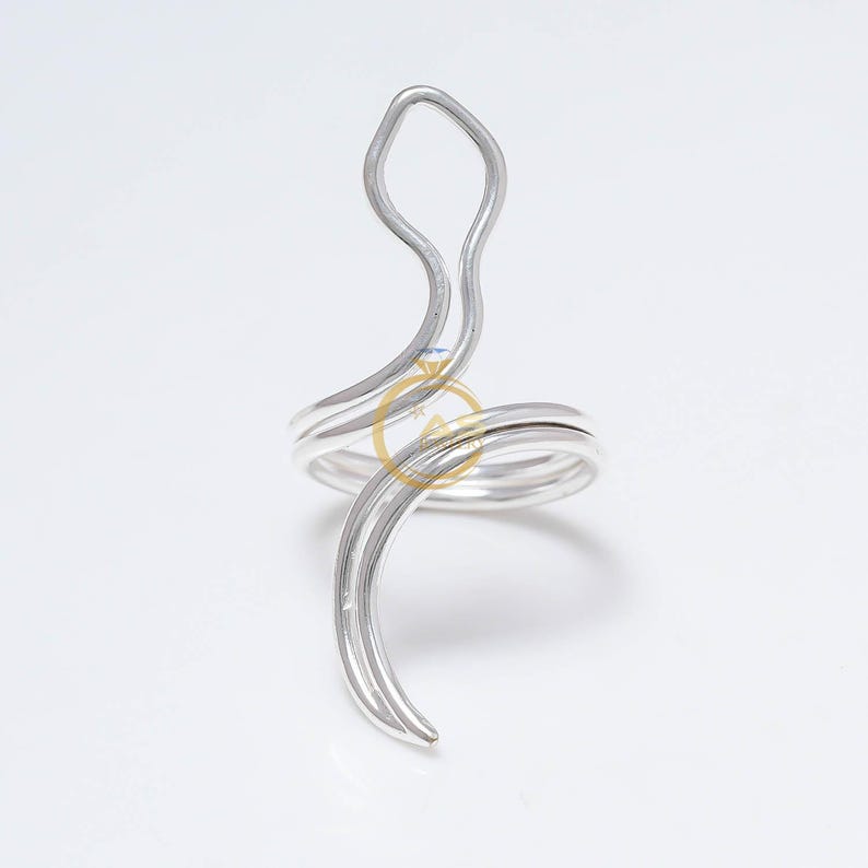 May include: A silver-colored ring with a unique, abstract design. The ring features a swirling band that wraps around the finger, with an elongated, curved element extending upwards. The ring is set against a white background.