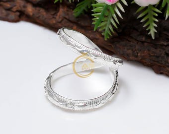 May include: Two silver rings with a detailed floral pattern. The rings are displayed on a white surface, with a dark brown wooden background and pink flowers in the upper right corner. The rings are likely for a wedding or engagement.