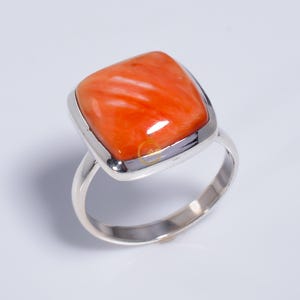 May include: A silver ring featuring a large, square-shaped, polished orange stone set in a smooth metal bezel. The stone displays natural swirling patterns and a glossy finish, while the band is a simple, polished silver metal loop.
