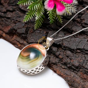 May include: A silver pendant necklace with an oval gemstone displaying brown, green, and white swirls. The gemstone is set in a decorative silver frame and suspended from a fine silver chain. The necklace is presented on a dark, textured background.