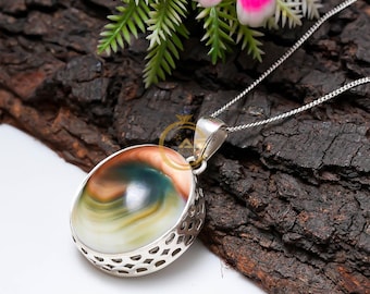 May include: A silver pendant necklace featuring an oval-shaped gemstone with swirling patterns of brown, green, and white. The gemstone is set in a decorative silver frame and hangs from a delicate silver chain. The necklace is displayed on a dark, textured surface.