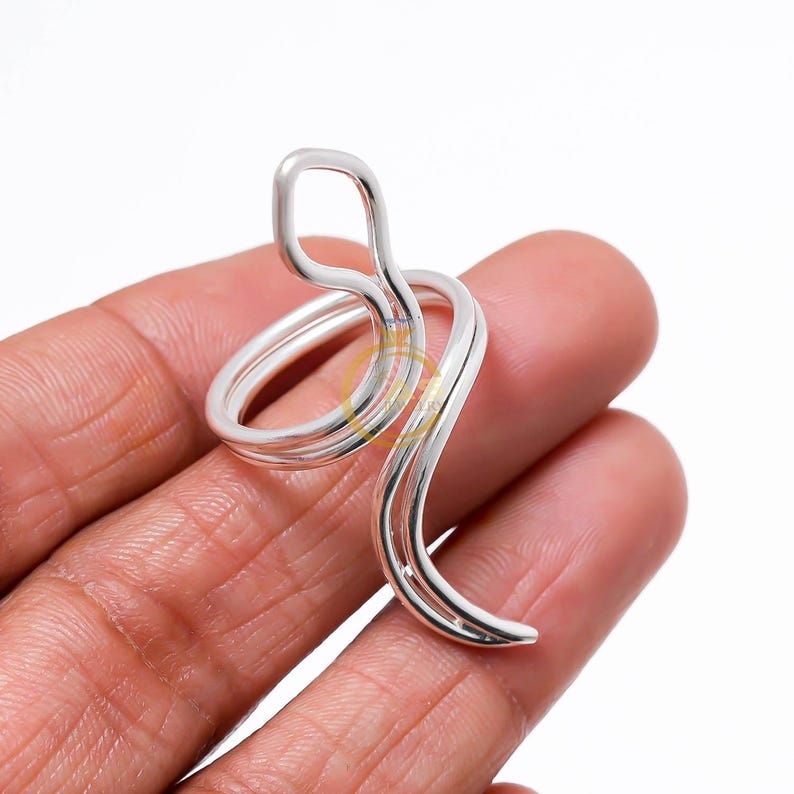 May include: A silver-colored ring with a unique, flowing design. The ring curves and loops, creating an abstract shape. The ring is made of a shiny, metallic material, and is shown on a person's finger.
