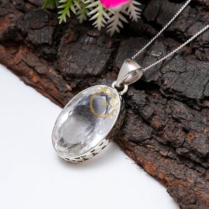 May include: A clear, oval-shaped crystal pendant with a silver setting and chain. The crystal is faceted, catching the light. The pendant hangs on a delicate silver chain. The background is a dark, textured surface.
