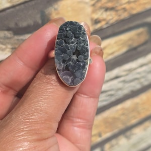 May include: A silver ring with an oval dark grey stone. The stone has a textured surface with a clustered, crystalline appearance, with hints of purple and blue. The ring is held in a hand, showing the jewellery's design and colour.
