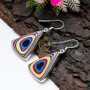 May include: Silver earrings with a triangular shape. Each earring has a layered pattern of black, blue, red, yellow, and white. The earrings are displayed on a dark, textured surface. The earrings are a striking piece of jewellery.