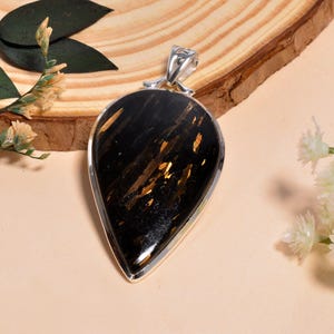 May include: A teardrop-shaped pendant with a black stone and gold flecks, set in a silver frame. The pendant hangs from a silver bail. The jewelry is displayed on a wooden surface with floral accents.