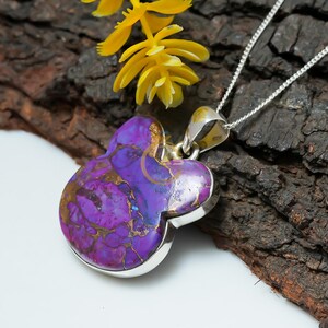 May include: A silver pendant necklace with a purple and gold clover-shaped stone. The stone has a marbled effect with gold veins. The necklace is presented on a dark wood surface with yellow flowers.