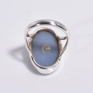 May include: A silver ring with a large oval blue agate stone. The ring is set in a silver band with a simple design. The back of the ring has the words "AS Jewelry" engraved on the stone.