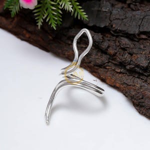 May include: A silver ring with an unusual, flowing design. The ring has a loop at the top and wraps around the finger. The jewellery is set against a dark wood background.