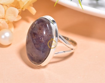 Può includere: Un anello d'argento con una grande pietra preziosa di forma ovale. La pietra preziosa è di colore viola scuro con macchie dorate e marroni. L'anello è incastonato in una semplice fascia d'argento.