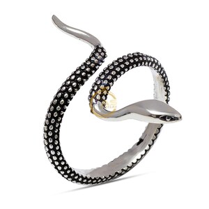 Puede incluir: Anillo de serpiente plateado con cuerpo texturizado negro y cabeza lisa y pulida. El anillo se enrolla, creando un diseño de serpiente. El cuerpo de la serpiente es negro con pequeños puntos en relieve. El anillo está sobre un fondo blanco.