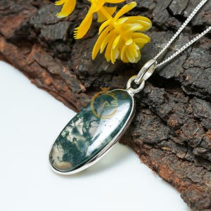 May include: A silver pendant necklace featuring an oval-shaped moss agate stone with green and white patterns. The stone is set in a silver bezel and hangs from a delicate silver chain. Yellow flowers and a dark wood background provide a natural backdrop.