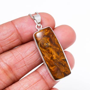 May include: A rectangular pendant with a silver-colored frame and a chain. The pendant features a polished, golden-brown stone with swirling patterns. The stone is set in a rectangular silver frame. The pendant is held by a hand.