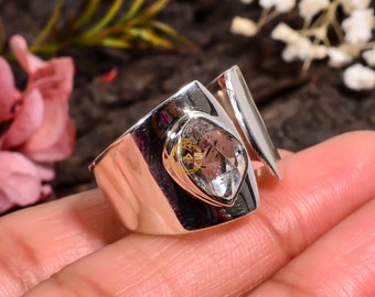 May include: A silver ring with a large, clear, faceted gemstone set in a silver band. The ring is open on one side and has a unique, modern design.
