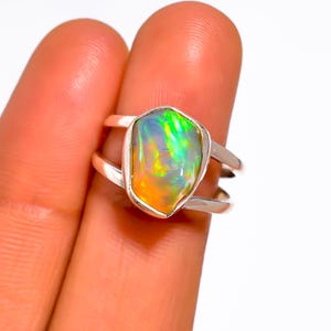 May include: A sterling silver ring featuring a vibrant, multi-colored opal gemstone. The opal displays a play of color with hues of green, yellow, and orange. The ring has a double-band design, showcasing the gemstone prominently.