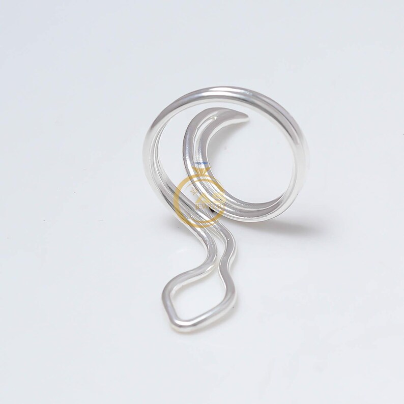 May include: A silver-colored, abstract metal ring with a looped design. The ring features a circular top and a wavy, elongated bottom section. The metal has a polished, reflective surface, and the design is set against a white background.