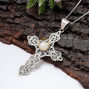 May include: A silver filigree cross pendant with a pearl center, suspended from a delicate chain. The cross features intricate openwork detailing and is displayed against a dark, textured background, showcasing the jewelry's craftsmanship.