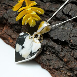 May include: A heart-shaped pendant with a black and white stone, set in a silver frame, hangs from a silver chain. A yellow flower rests near the pendant. The pendant's stone has a unique pattern.