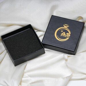 May include: Black jewelry box with a gold logo that reads "AS JEWELRY". The box is open, revealing a black foam insert. The box is on a white, textured surface.