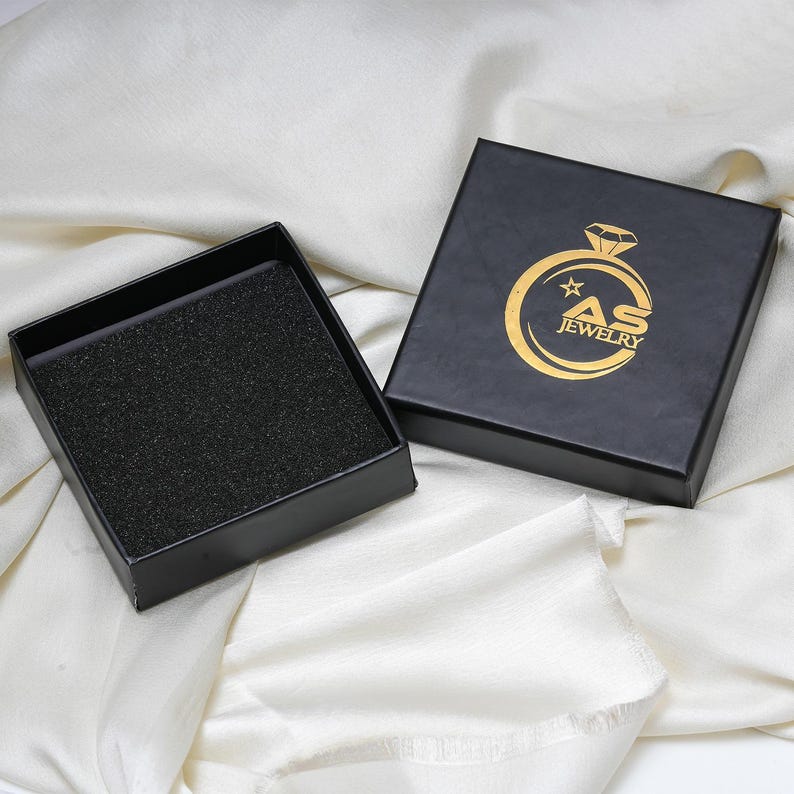May include: Black jewelry box with a gold logo that reads "AS JEWELRY". The box is open, revealing a black foam insert. The box is on a white, textured surface.