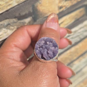 May include: A silver ring featuring a large, round, lavender-colored druzy stone. The stone has a bubbly, textured surface and is set in a simple silver band. The ring is shown on a finger.