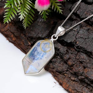 May include: A silver necklace featuring a geometric pendant with a rainbow moonstone. The pendant is a clear, multifaceted crystal with blue and white internal reflections. The necklace is displayed on a dark, textured wooden surface.