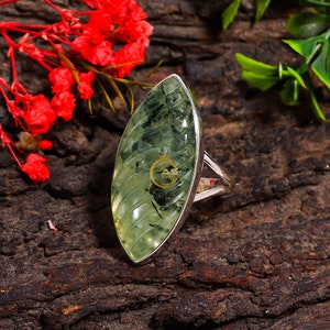 Puede incluir: Un anillo de plata con una piedra preciosa ovalada de color verde. La piedra preciosa tiene un patr&oacute;n estriado y un centro verde oscuro.