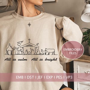 Może przedstawiać: Beżowa bluza z czarnym haftem przedstawiającym scenę narodzin z tekstem "All is calm All is bright". Na bluzie jest również wyszyty tekst "EMB I DST I JEF I EXP I PES I VP3".