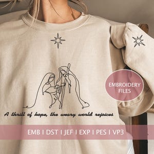 以下が含まれることがあります： ベージュのトレーナーに、黒い線で描かれたキリスト降誕のシーンと「A thrill of hope, the weary world rejoices」というテキストが書かれています。「EMB I DST I JEF I EXP I PES I VP3」というテキストもトレーナーに書かれています。このトレーナーは刺繍用です。