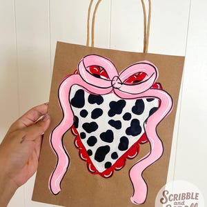 May include: Brown paper gift bag with a hand-painted design of a pink bow and a cow-print bandana. The bag has brown paper handles and the text "Scribble and Scroll by Chio" in the bottom right corner.