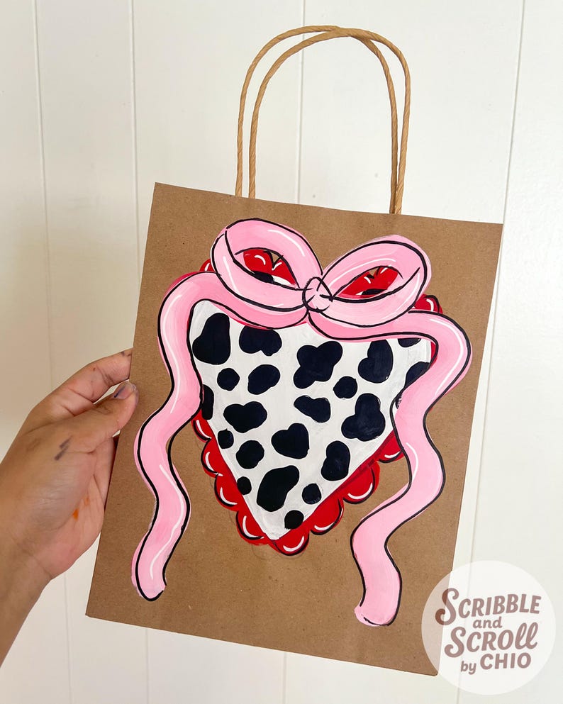 May include: Brown paper gift bag with a hand-painted design of a pink bow and a cow-print bandana. The bag has brown paper handles and the text "Scribble and Scroll by Chio" in the bottom right corner.