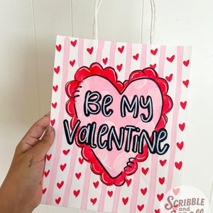 May include: A Valentine's Day gift bag with a white background and pink vertical stripes. Red hearts are scattered across the bag. A large pink heart with a red scalloped border and the words "Be My Valentine" is the focal point.