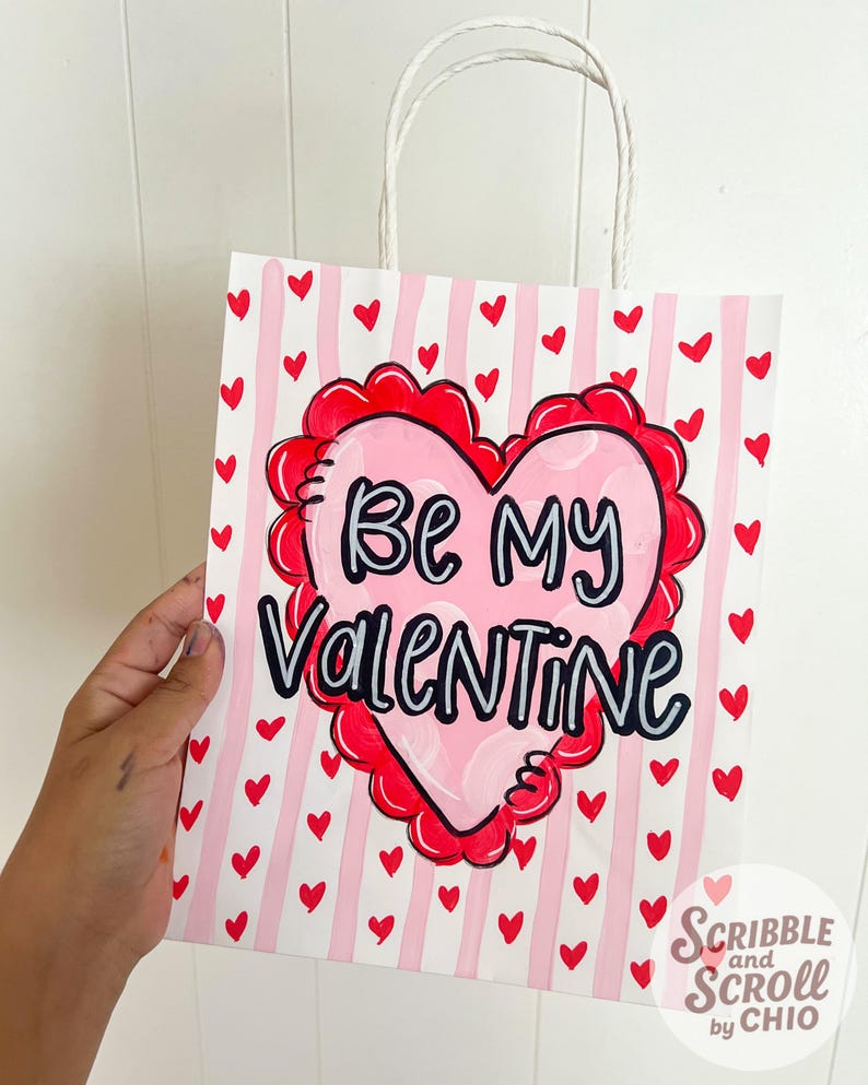 May include: A Valentine's Day gift bag with a white background and pink vertical stripes. Red hearts are scattered across the bag. A large pink heart with a red scalloped border and the words "Be My Valentine" is the focal point.