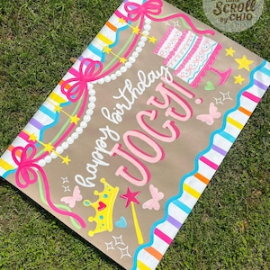 May include: A rectangular birthday sign on brown paper featuring the text "happy birthday JOCY!" in pink and white script. The design includes a pink cake, a yellow crown, a star wand, butterflies, and colorful striped borders with pink bows.