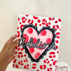 May include: White gift bag with a red leopard print pattern. A pink heart with red polka dots and the name "Adelina" in cursive is centered on the bag. The heart is outlined in black. White paper handles.