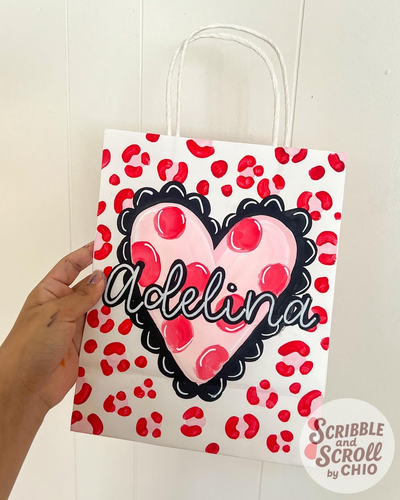 May include: White gift bag with a red leopard print pattern. A pink heart with red polka dots and the name "Adelina" in cursive is centered on the bag. The heart is outlined in black. White paper handles.