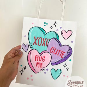 May include: White paper gift bag with a hand-painted design of heart-shaped candies. The bag features the phrases "XOXO", "Cutie", and "Hug Me" in red lettering. Additional hearts and decorative elements are scattered around the bag.