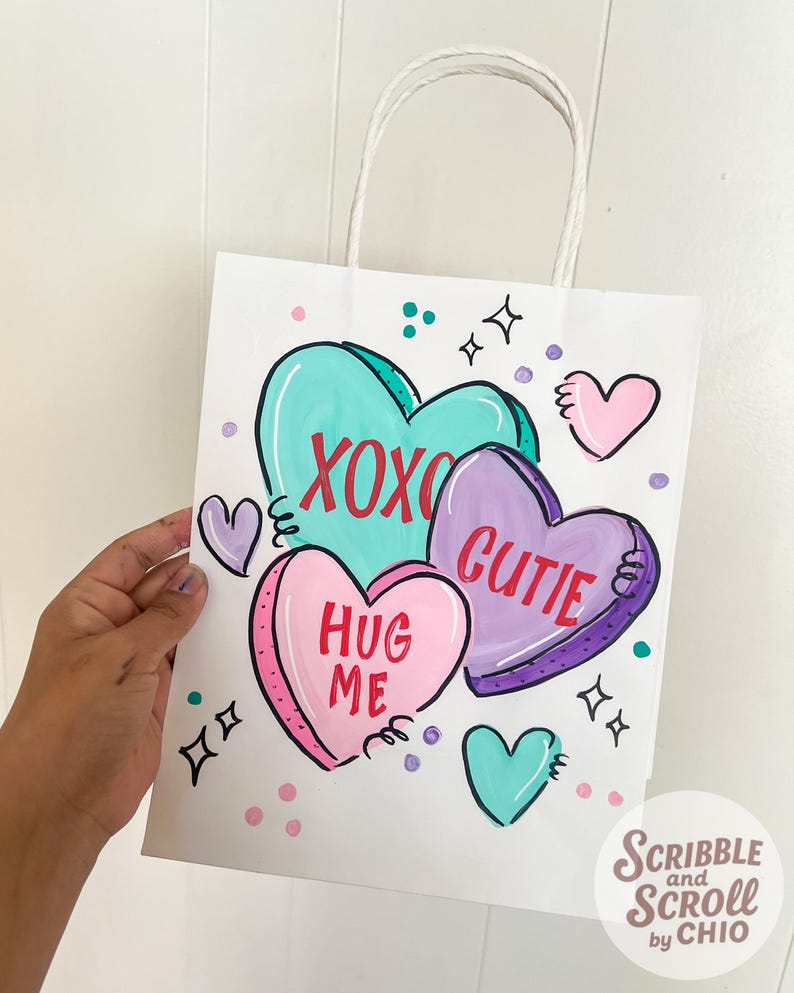 May include: White paper gift bag with a hand-painted design of heart-shaped candies. The bag features the phrases "XOXO", "Cutie", and "Hug Me" in red lettering. Additional hearts and decorative elements are scattered around the bag.