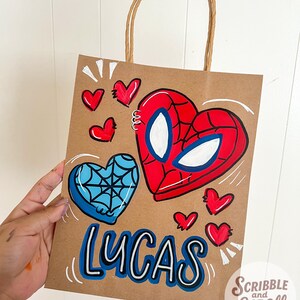 May include: A brown paper gift bag with a brown twisted paper handle. The bag features a hand-painted design of a red and blue heart with a spiderweb design and the name "LUCAS" in blue and white. Small red hearts are scattered around the design.