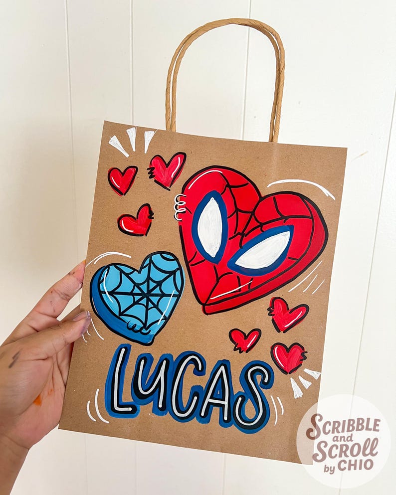 May include: A brown paper gift bag with a brown twisted paper handle. The bag features a hand-painted design of a red and blue heart with a spiderweb design and the name "LUCAS" in blue and white. Small red hearts are scattered around the design.