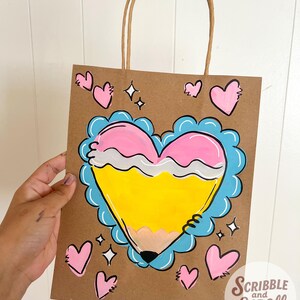 May include: A brown paper gift bag with a hand-painted design. The design features a yellow pencil shaped like a heart, outlined in blue and pink, with additional pink hearts and sparkles. The bag has a brown paper handle and the text "Scribble and Scroll by Chio".