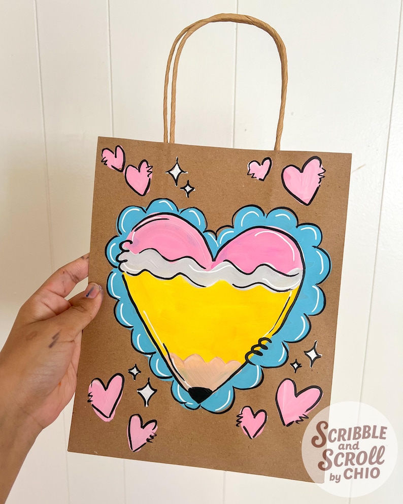 May include: A brown paper gift bag with a hand-painted design. The design features a yellow pencil shaped like a heart, outlined in blue and pink, with additional pink hearts and sparkles. The bag has a brown paper handle and the text "Scribble and Scroll by Chio".