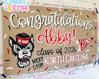 May include: A rectangular tan sign featuring the hand-painted text Congratulations Abby! class of 2026 next stop... NORTH CAROLINA! with a red and white GO PACK pennant, an NC State wolf mascot graphic, and scattered red, white, and black confetti.