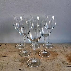 Può includere: Un gruppo di sei calici da vino trasparenti. I calici presentano un design classico con una coppa arrotondata, un gambo sottile e una base circolare. I gambi hanno un elemento decorativo. I calici sono disposti su una superficie di legno.