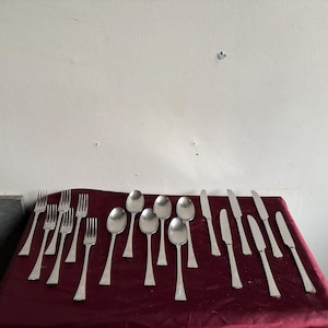 Può includere: Un set di posate, tra cui forchette, cucchiai e coltelli, disposti su un panno rosso bordeaux. Le posate sono in acciaio inossidabile lucido.