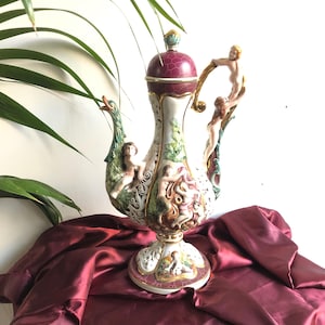 Può includere: Una grande e ornata teiera in ceramica con un coperchio color borgogna e un corpo bianco. La teiera è decorata con dettagli intricati, tra cui figure di persone e un uccello. La teiera è appoggiata su una superficie di tessuto rosso.