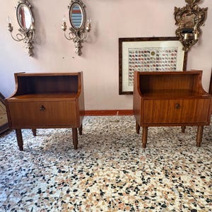 May include: A pair of matching wooden bedside tables, each featuring an open top shelf and a single lower drawer with a small circular metal pull. The bedside tables are supported by four cylindrical legs and have a smooth, medium-toned wood grain finish.