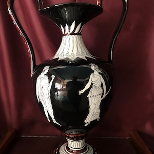 Può includere: Vaso in ceramica nero e bianco con due manici e un design decorativo con figure in bianco su sfondo nero. La base del vaso è decorata con motivi rossi e bianchi.